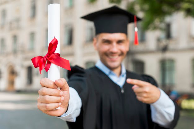 homem sorridente trajando vestes de formatura com um diploma na mão